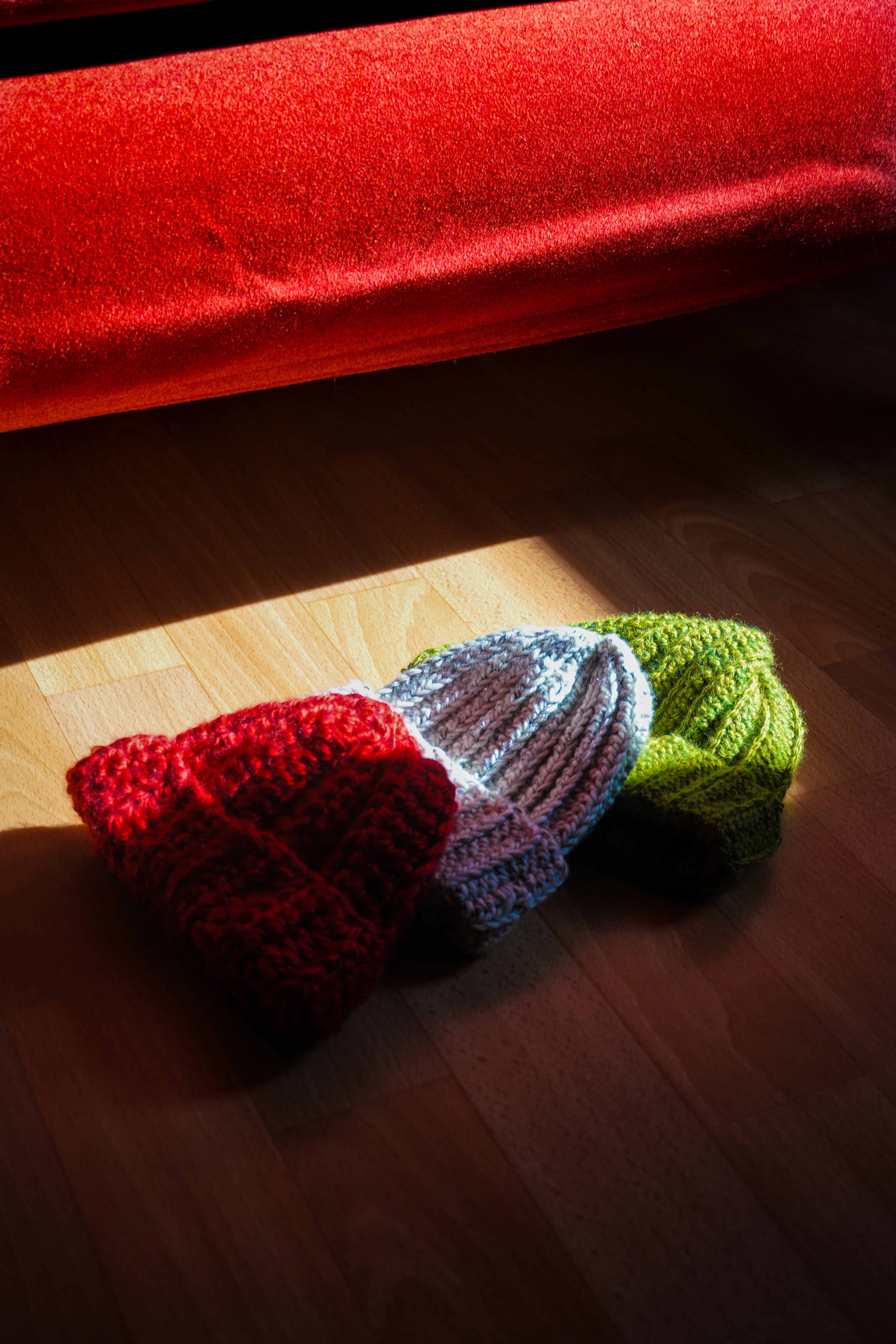 Three crocheted hats in red, blue, and green on a wooden floor with a red blanket in the background.